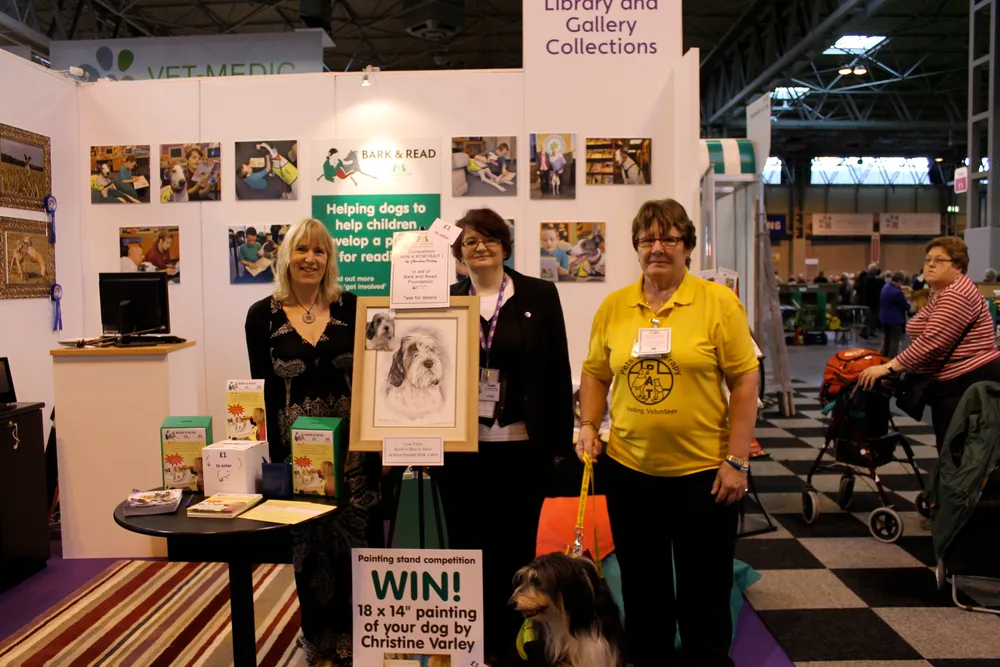 Christine and two Kennel Club volunteers with a dog on the Bark & Read stand at Crufts, beside a competition poster to win a painting of your dog