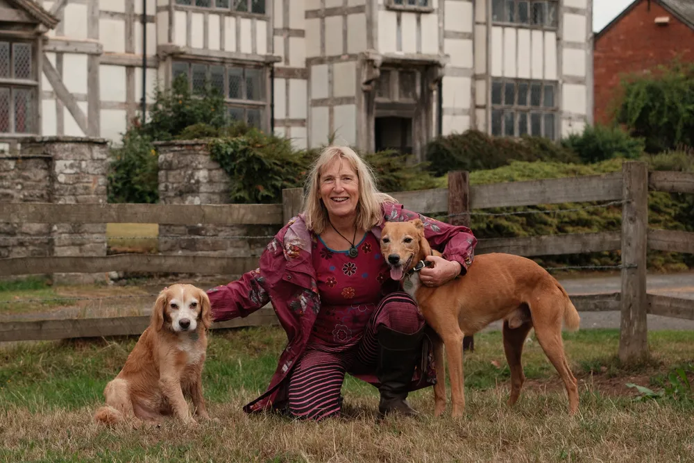 Christine crouching on a lawn between two dogs, in front of a black and white timber-framed house