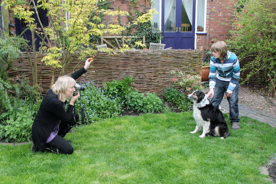 Christine kneeling in a garden with a camera, holding up a treat, while a boy holds a collie still