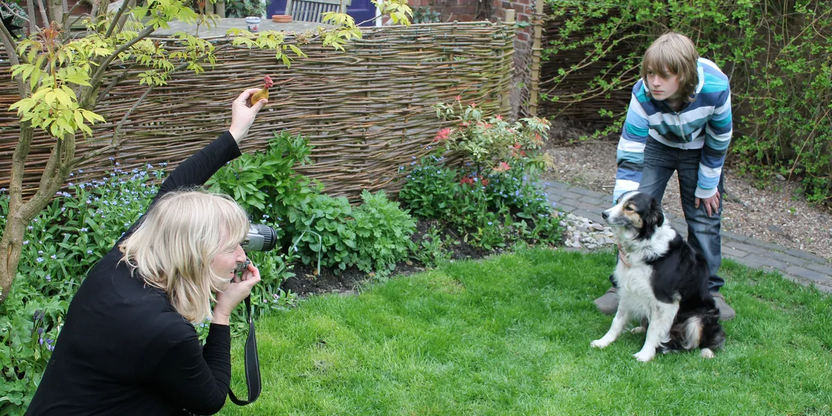Christine crouching in a garden with her camera, photographing a black and white collie while a boy holds its attention